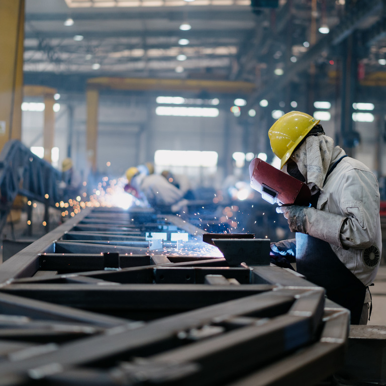 Workers are engaged in welding activities, fabricating metal components while sparks fly in a well-lit factory environment during the day.