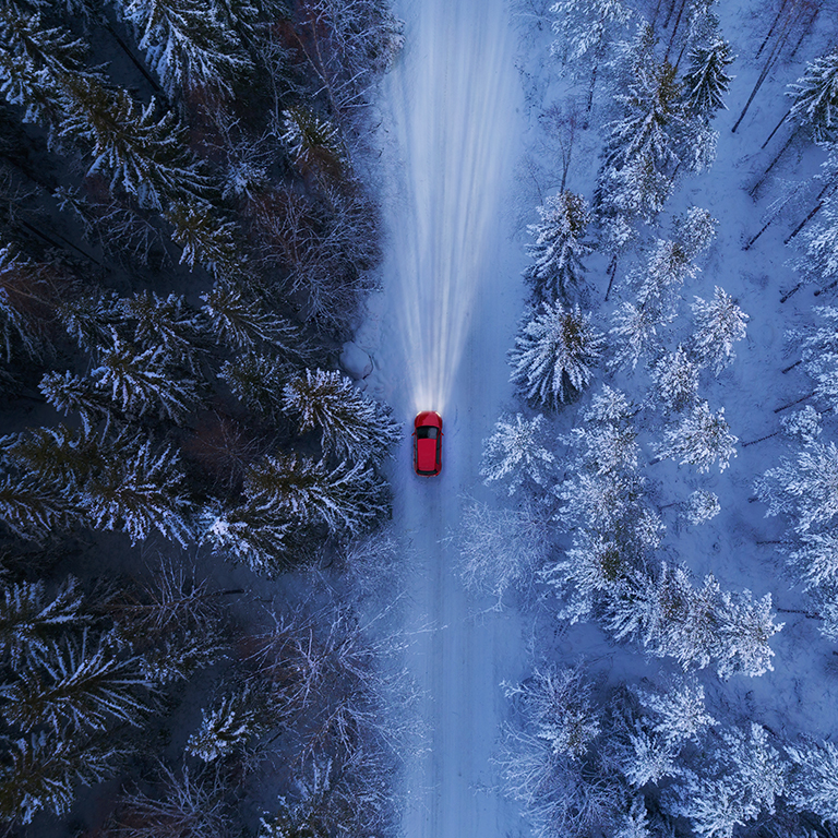Birdseye view of a road through a forest in winter with red car driving with headlights on