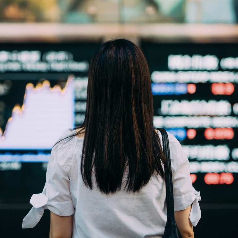A woman stands in front of a stock market display. She observes the changing data on stock prices and market trends. The setting is a busy financial center during business hours.