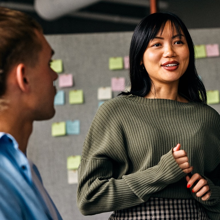 A woman stands in front of a group during a meeting at an office. She talks while a man listens. Sticky notes are on the wall behind them.