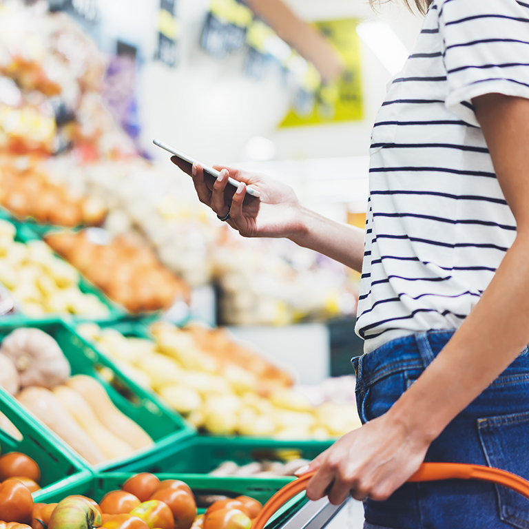 Young woman shopping healthy food in supermarket blur background. Female hands buy nature products using smart phone in store. Hipster at grocery using mobile. Person comparing price of produce