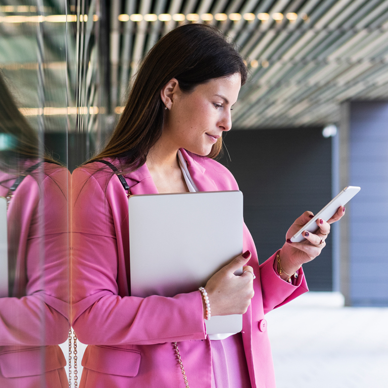 Female young entrepreneur with laptop using mobile phone while leaning on glass wall