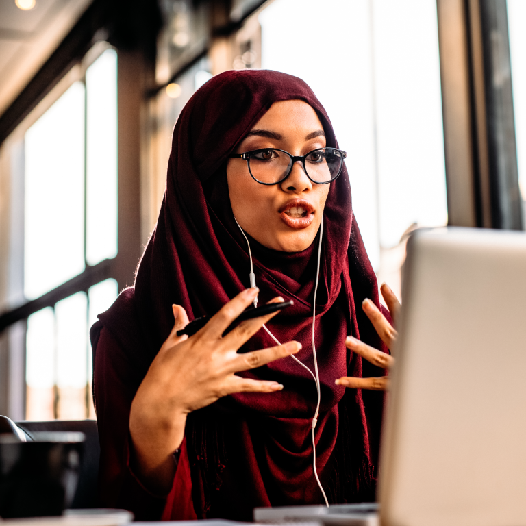 Young woman in hijab talks during online meeting in cafe using a laptop.